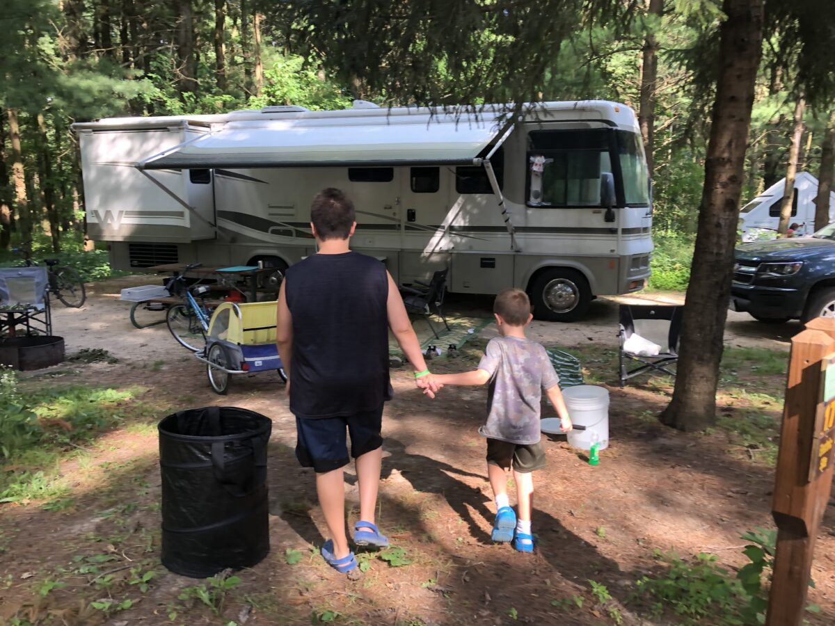 Two boys walking hand in hand toward an RV at a wooded campsite